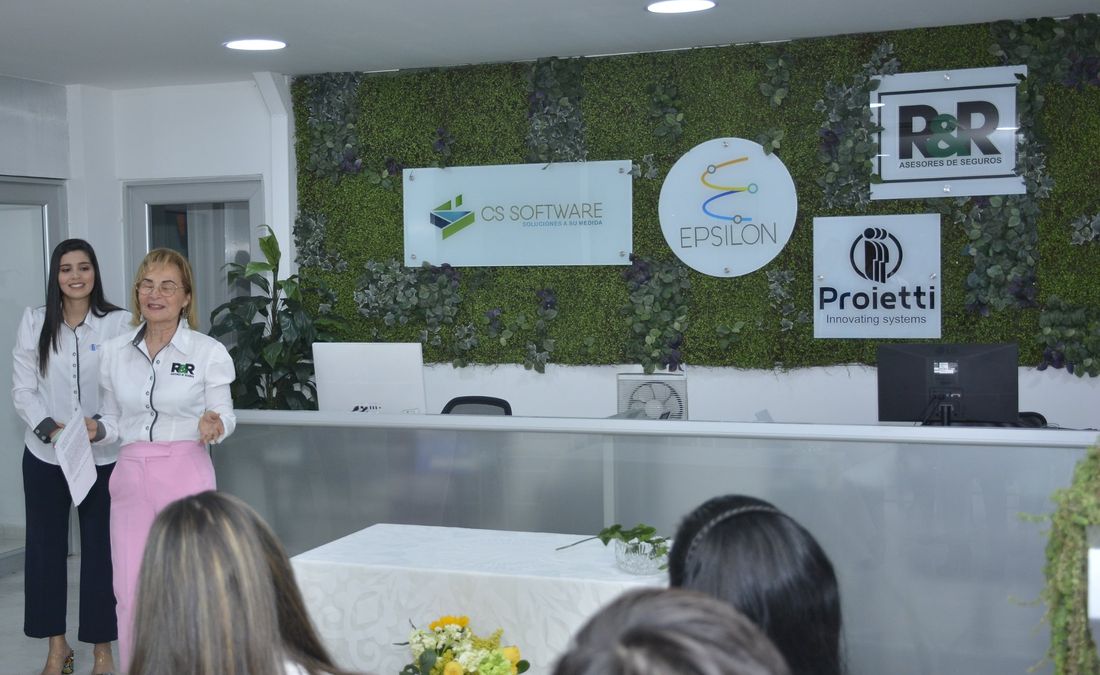 Inauguración de la oficina en 2022: dos personas hablan al público frente a la pared con los logos de las empresas del grupo y el mostrador de atención.